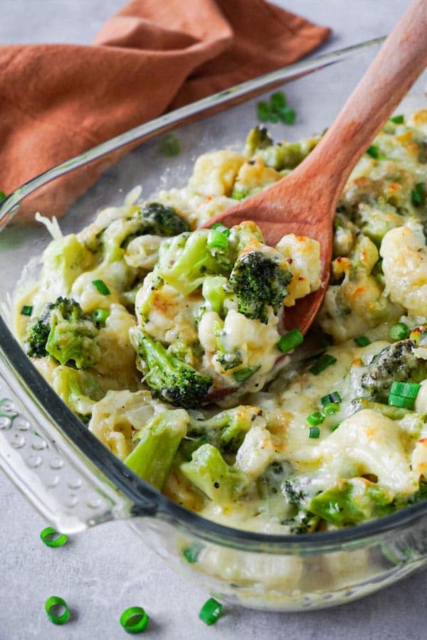 Wooden spoon scooping out creamy and cheesy cauliflower with broccoli from glass baking dish.