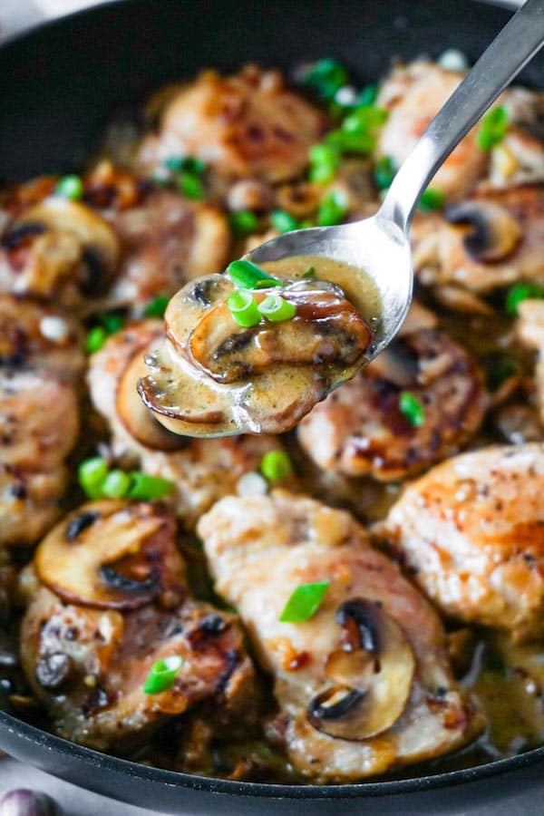 A silver spoon with creamy mushroom sauce garnished with chopped green onions with fried chicken thighs on the background.