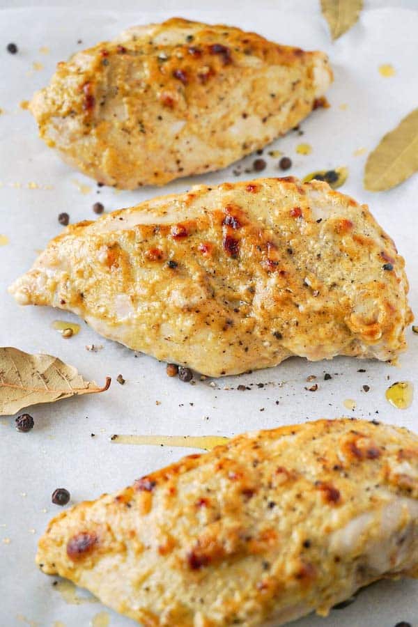 3 baked Dijon mustard chicken breasts drizzled with olive oil lying on a white parchment paper with some bay leaves and pre-grounded black pepper.
