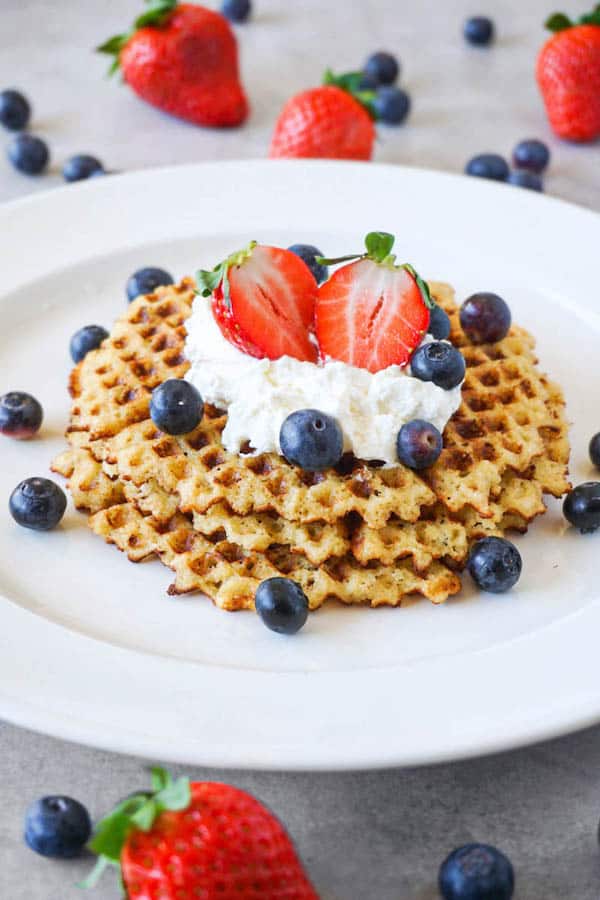 Crispy waffles topped with whipped cream, fresh strawberries and blueberries on a large white ceramic plate standing on a grey cooking top, fresh berries lying arround.