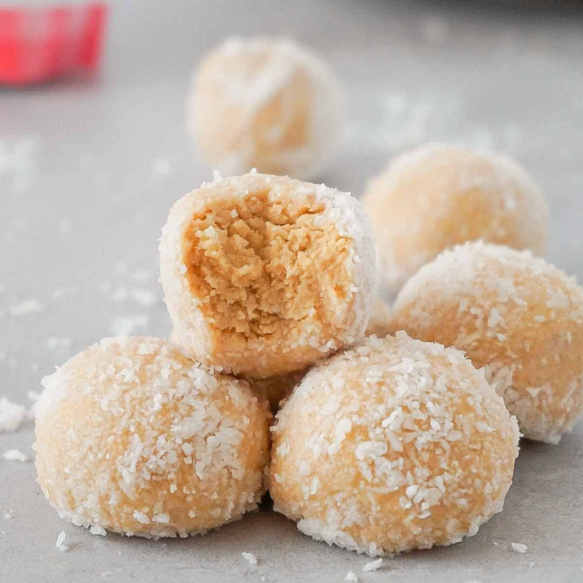 Pyramid of keto peanut butter balls covered with coconut flakes on a grey cooking surface, one bite is taken from the top one.