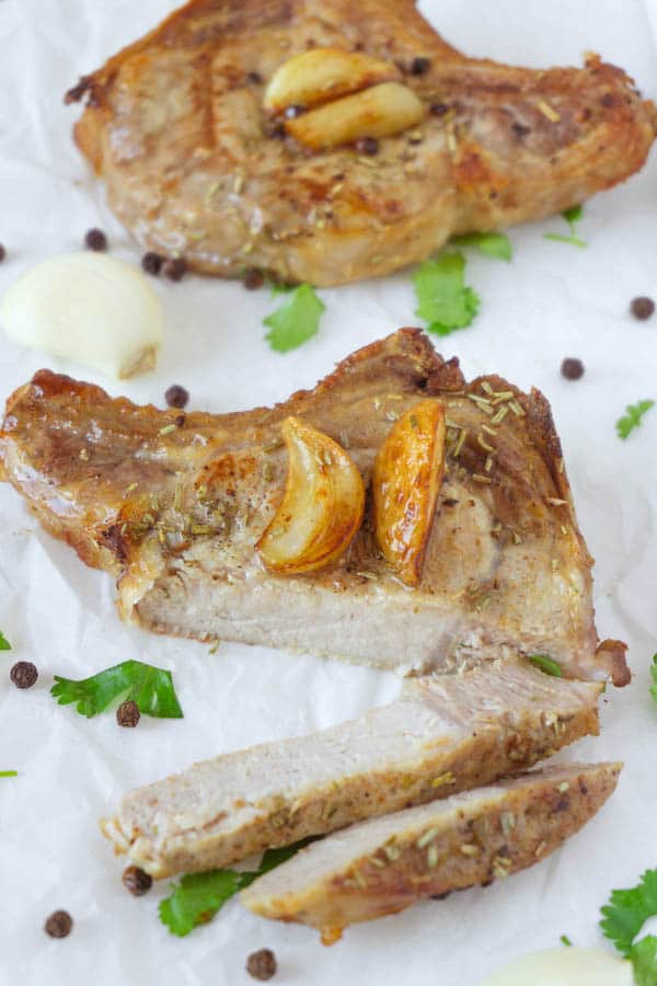 Two baked pork chops garnished with baked garlic on a white parchment paper, one pork chop is sliced.