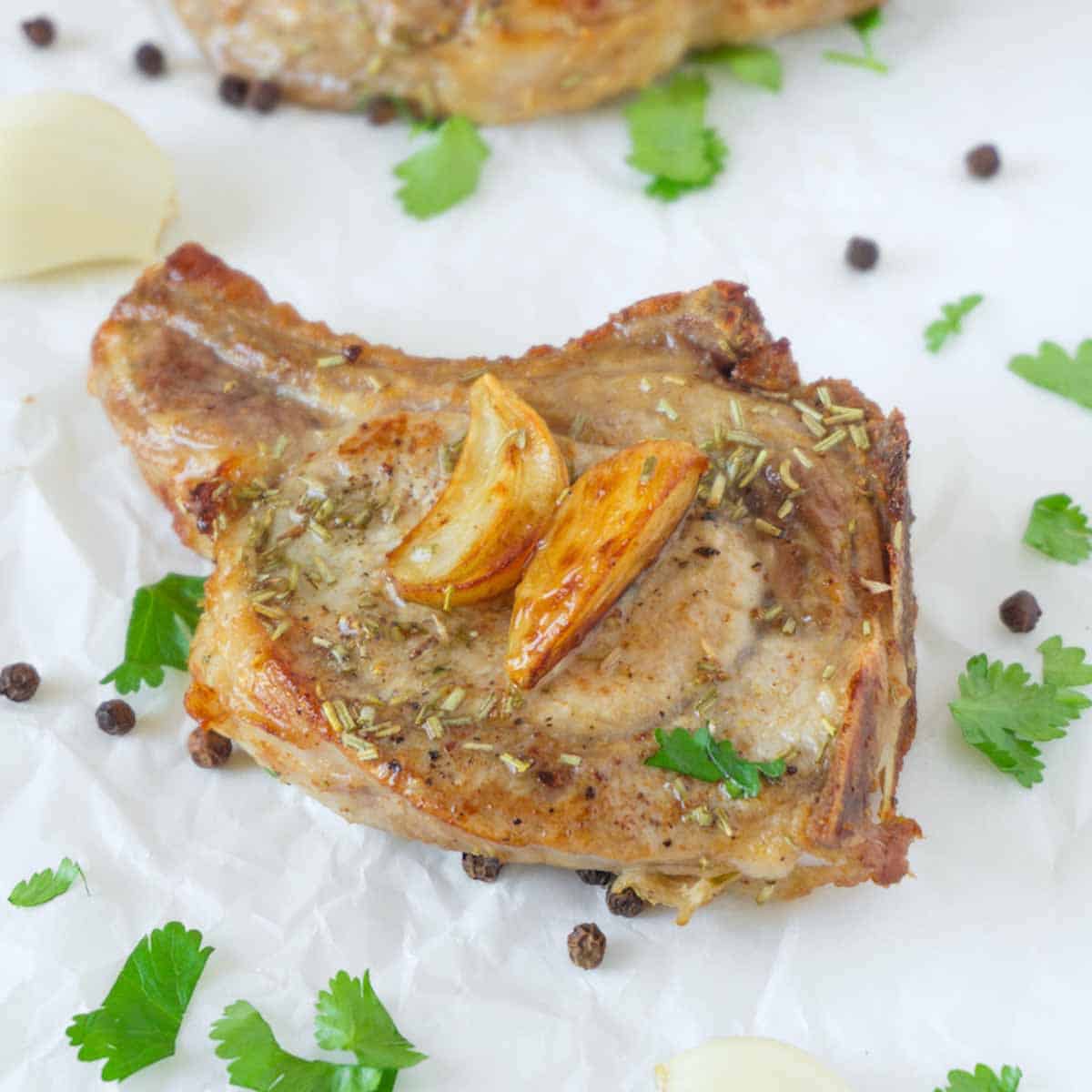 Close up picture of baked pork chop garnished with baked garlic, pieces of freshly-chopped cilantro and pre-ground black pepper are lying around.