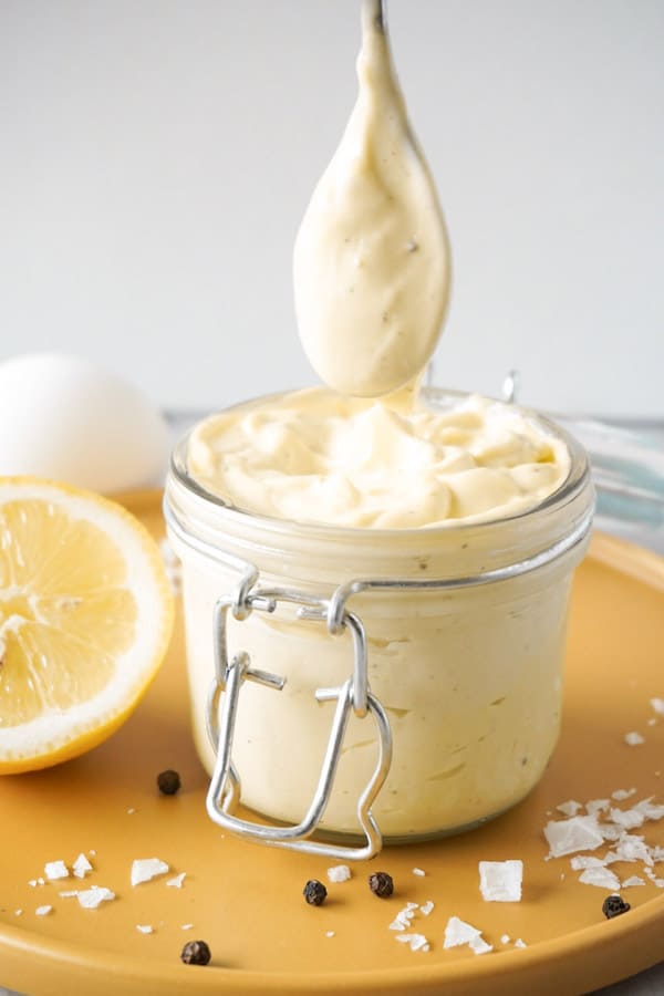 Homemade mayonnaise in a glass jar standing on a yellow plate with some lemon, salt and black pepper lying around.