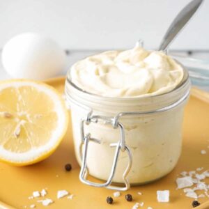 Close up picture of keto mayonnaise in a glass jar with a silver spoon inside standing on a yellow plate with some lemon, salt and black pepper lying around.