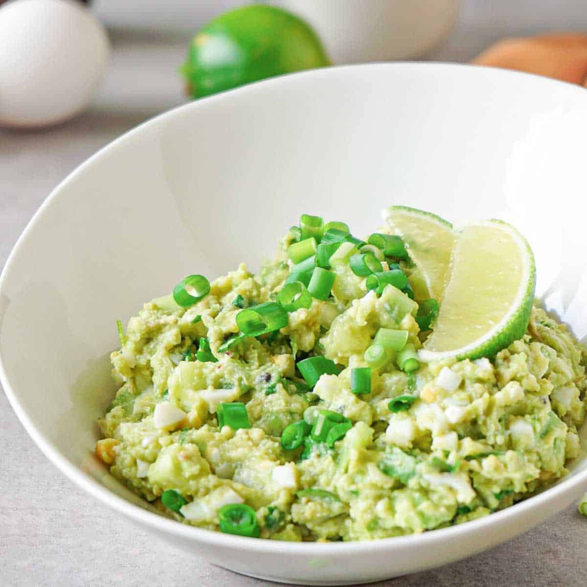 A white porcelain bowl with avocado egg salad garnished with chopped green onions and lime wedges.
