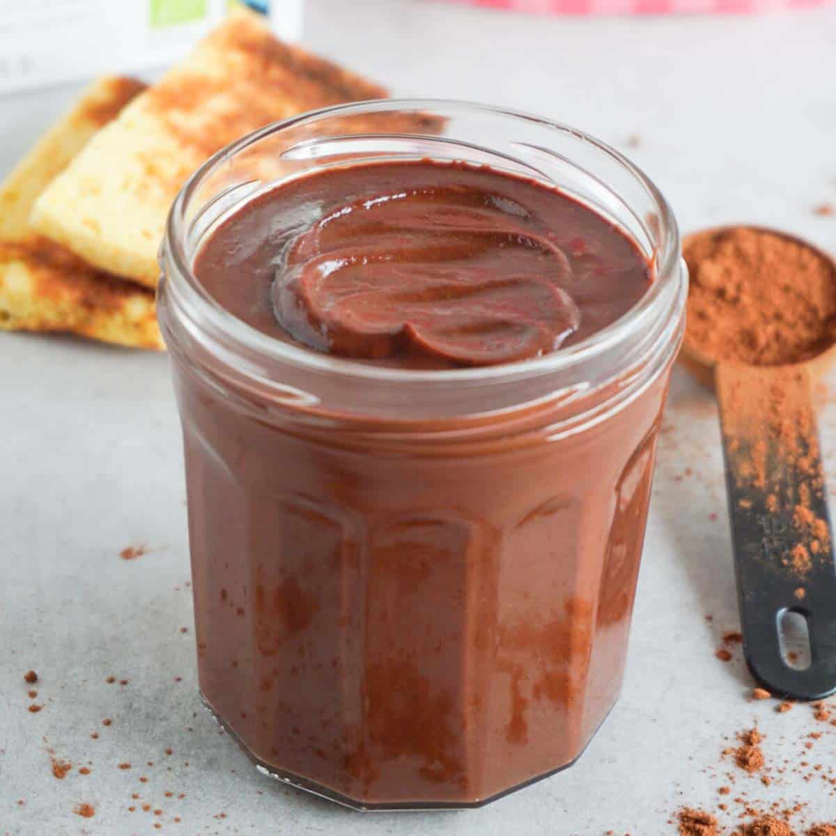 Close up picture of chocolate spread in an opened glass jar on a grey cooking surface with cocoa powder sprinkled on it.