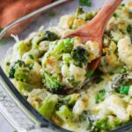 Close up of a wooden spoon scooping out creamy cauliflower with broccoli from a glass baking dish.