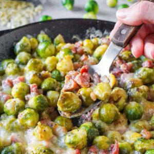 Cast iron skillet full of cheesy brussels sprouts with bacon with a silver spoon digging in.