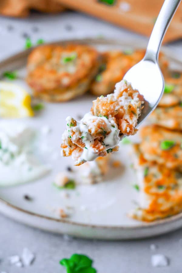 Piece of salmon cake on a silver fork with some full-fat sour cream, plate of salmon cakes in the background.