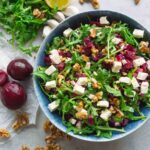 Top down picture of a blue ceramic bowl full of arugula red beet salad with feta and walnuts on a grey working top, whole beets, garlic, arugula leaves, walnuts are lying around.