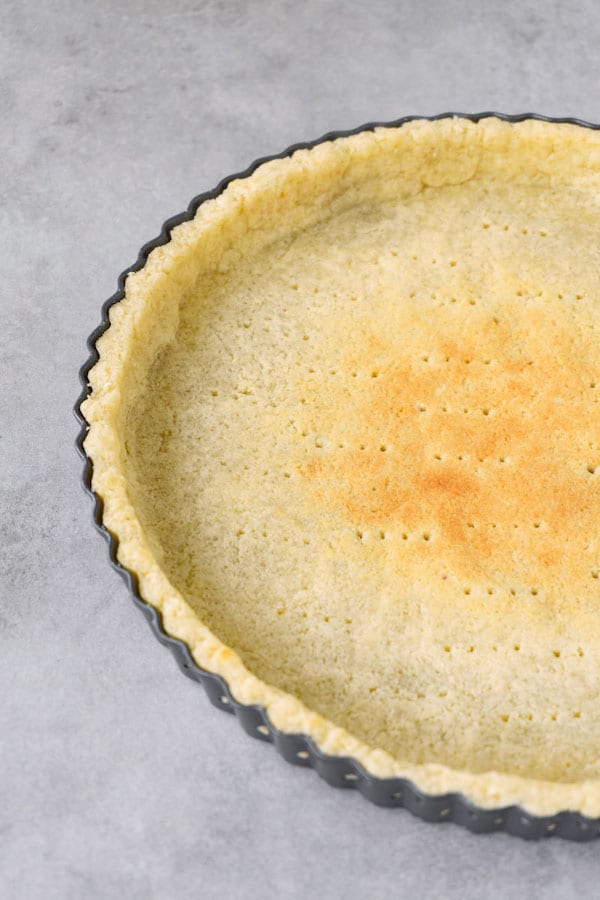 Partially blind baked gluten-free pie crust, the base pricked with a fork, in a tart pan lying on a grey working surface.