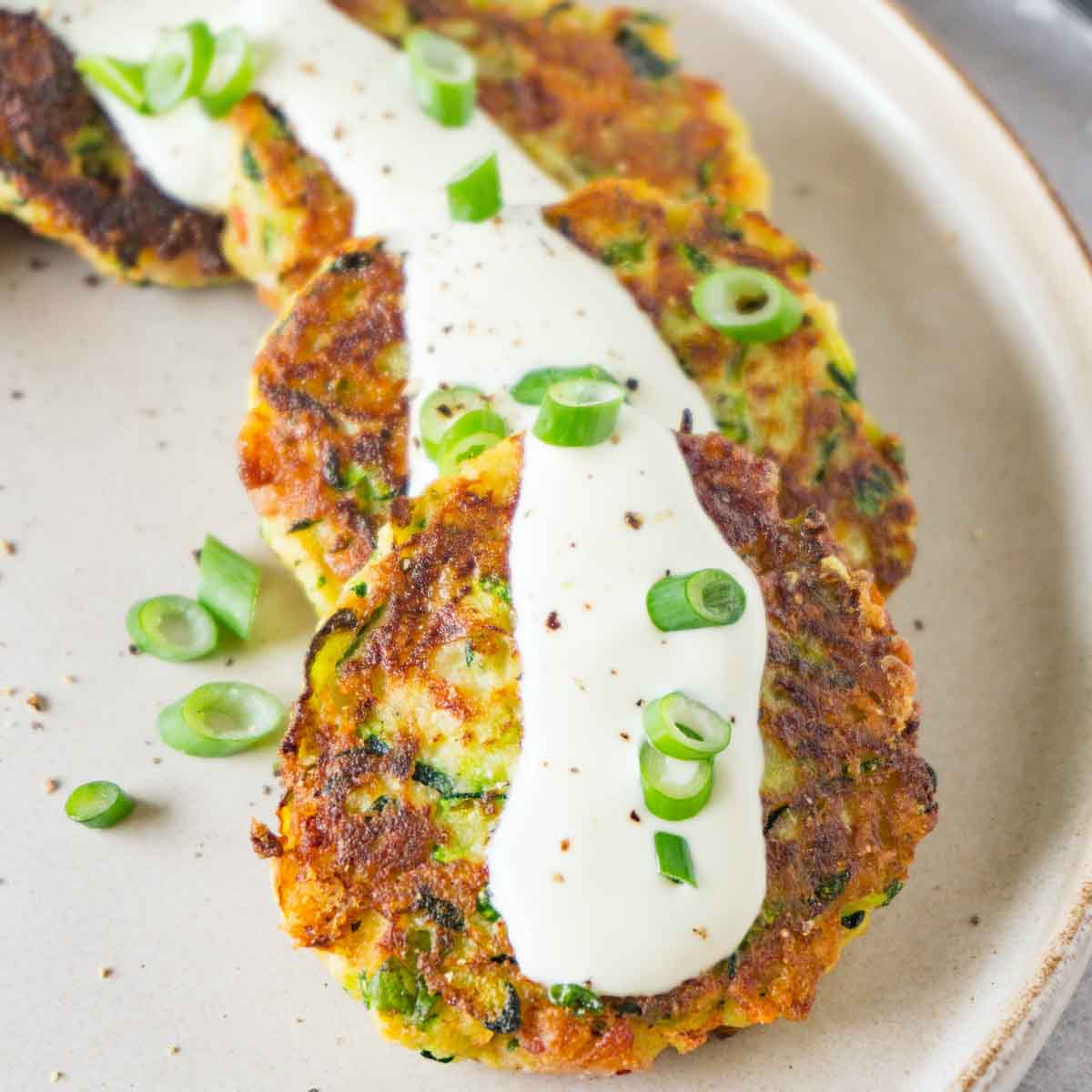 Close up shot of zucchini fritters on a round beige plate garnished with sour cream, chopped green onions and ground black pepper.