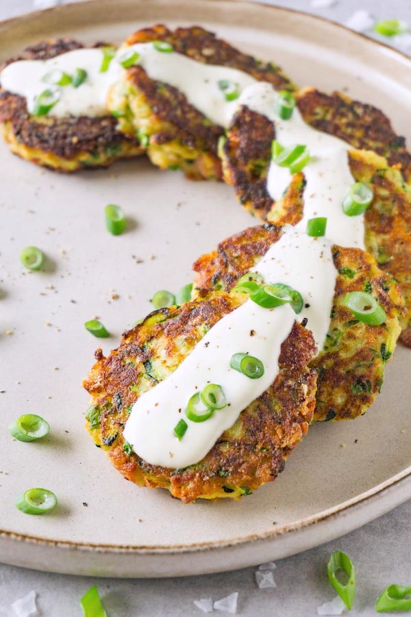 Freshly made fritters with zucchini on a round ceramic plate garnished with sour cream and chopped green onions and freshly ground black pepper.