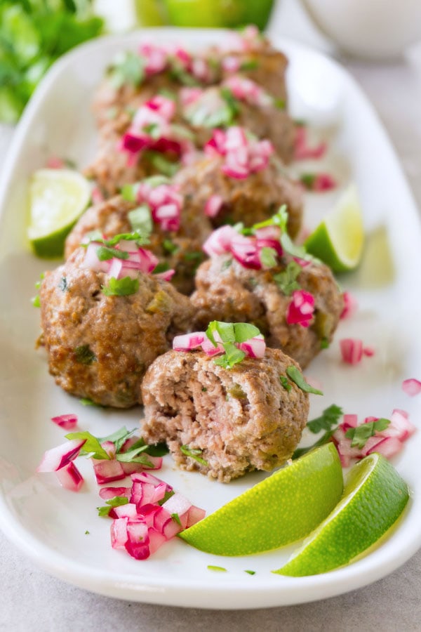 Beef meatballs topped with red onions and chopped cilantro, served on a long, white plate with lime wedges, bite taken from one meatball.