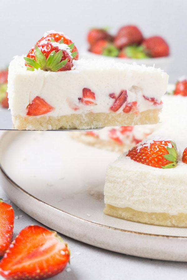 A slice of a no bake keto strawberry cheesecake decorated with fresh strawberries and shredded coconut on a cake server, the rest of the cake is above the slice.