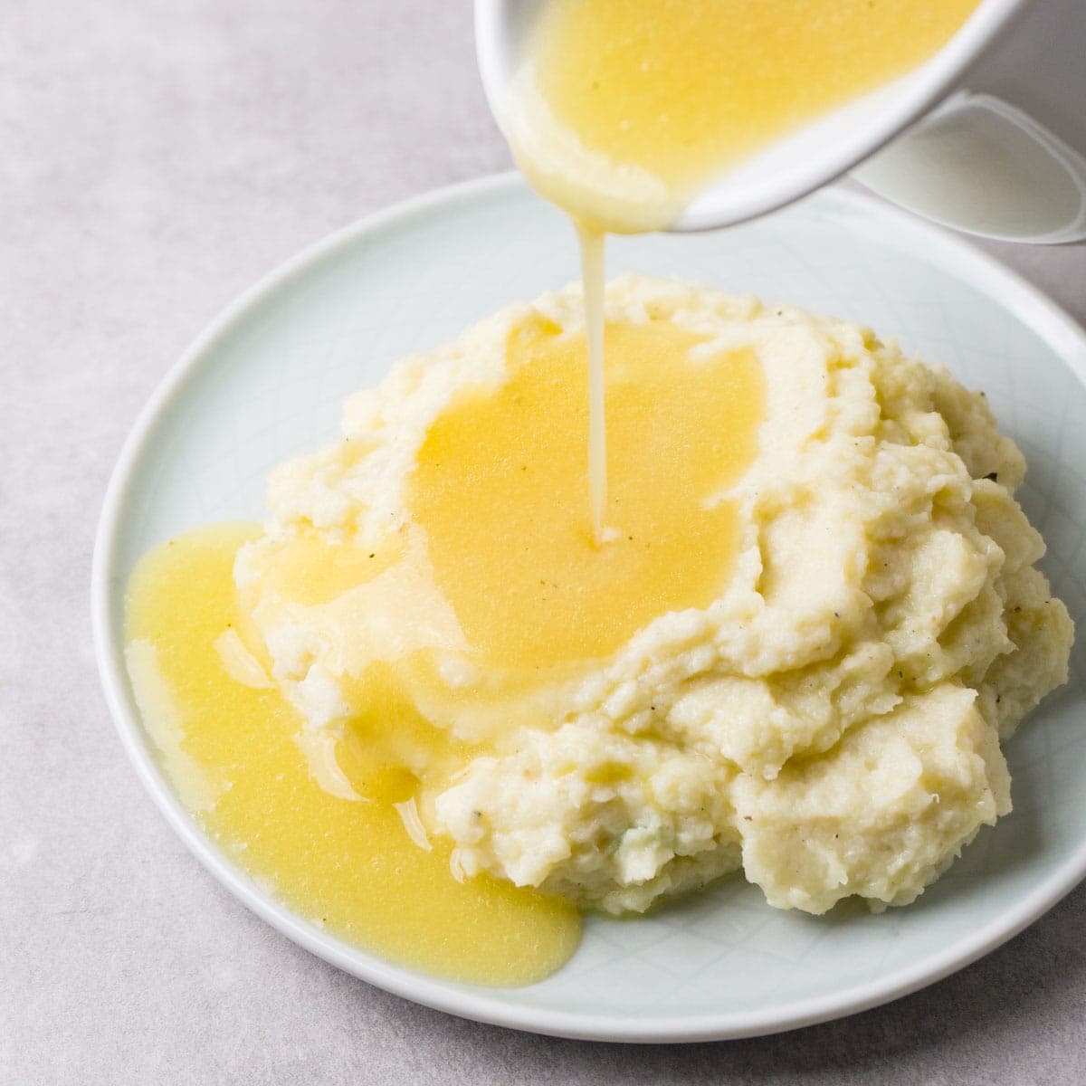 Close up shot of a gravy sauce pouring from a white sauce jug on top of cauliflower mash on a small round plate.
