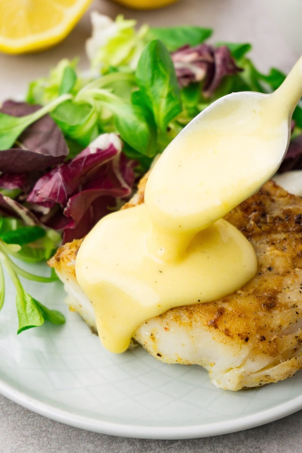 Pouring hollandaise sauce from a spoon on top of a piece of fried cod with fresh green salad as a side dish.