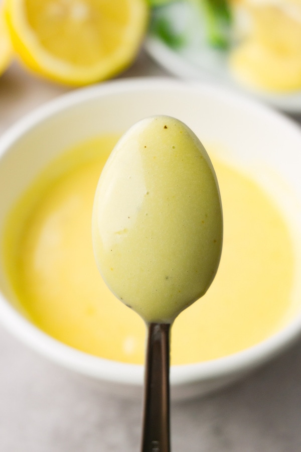 A spoon coated with freshly-made hollandaise sauce, a white sauce bowl is on the background.