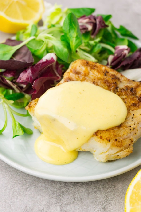 A piece of fried cod topped with hollandaise with a green salad on the side served on a small round plate.