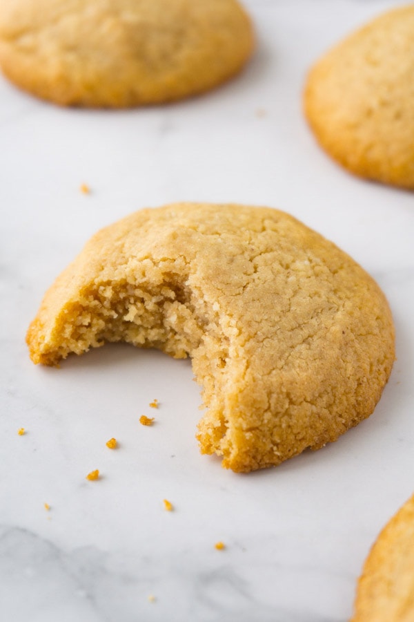 Peanut butter cookie lying on a white marble surface, one bite taken, crumbs and more cookies are lying around.