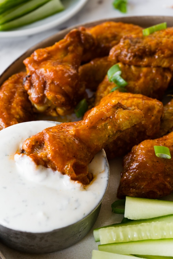 Buffalo wings on a plate with cucumber sticks and a small bowl filled with ranch dip, one drumettes is lying in the ranch dip bowl.