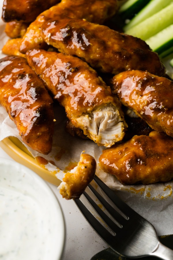 Buffalo chicken tenderloins served on a plate with ranch dip, one piece taken with a fork from one tenderloin, cucumber sticks on the background.