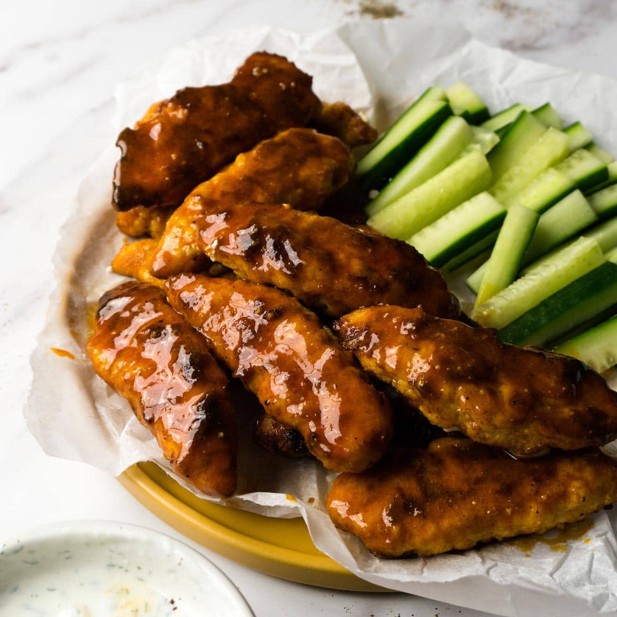 Buffalo chicken tenders on a plate lined with parchment paper, cucumber sticks on the back of the plate and ranch dip on the front.
