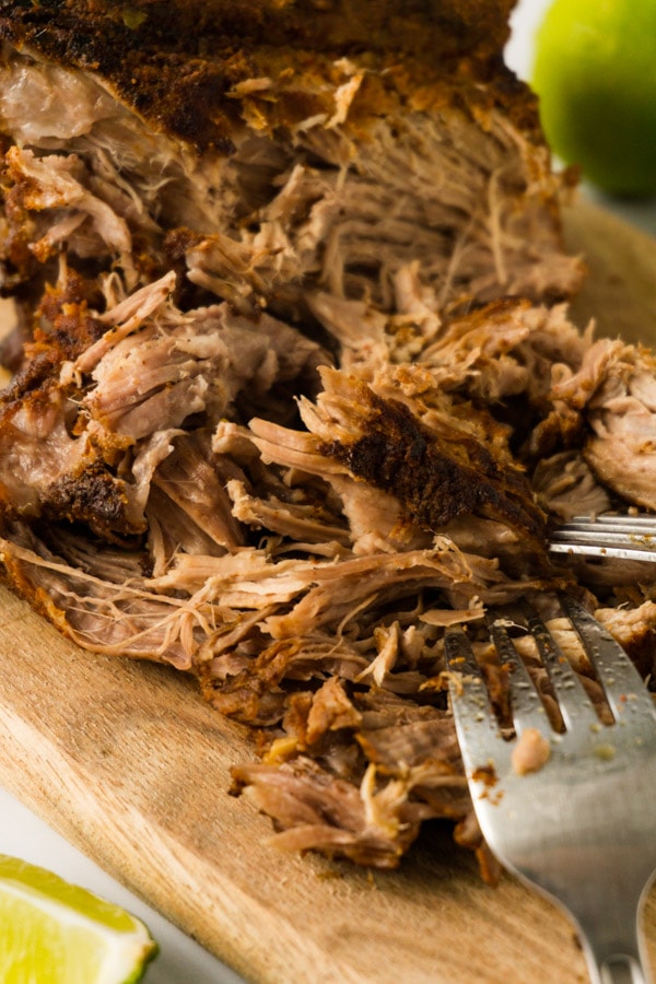 A piece of slow-cooked pork butt on a wooden board, half of the pieces shredded using two forks that are lying near.