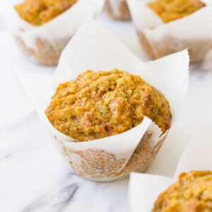 Close up shot of a carrot cake muffin in a muffin liner standing on a marble surface, more muffins are lying around.