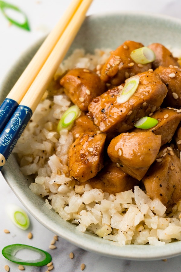 A small bowl with cauliflower rice and teriyaki chicken garnished with sesame seeds and freshly chopped green onions, chopsticks are lying on the side of the bowl.