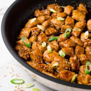 Close up shot of a skillet filled with teriyaki chicken topped with sesame seeds and green onions.
