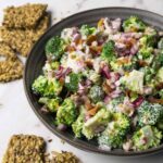 A bowl full of broccoli salad with creamy dressing, bacon bits and red onions, seed crackers lying near the bowl.