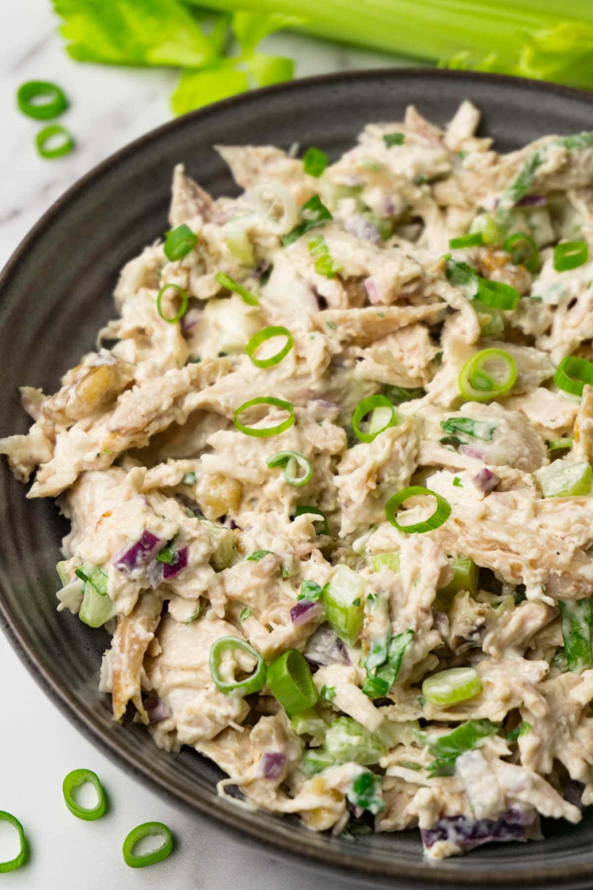 Close up shot of a salad with shredded chicken, red onions, celery and creamy dressing, topped with chopped spring onions.