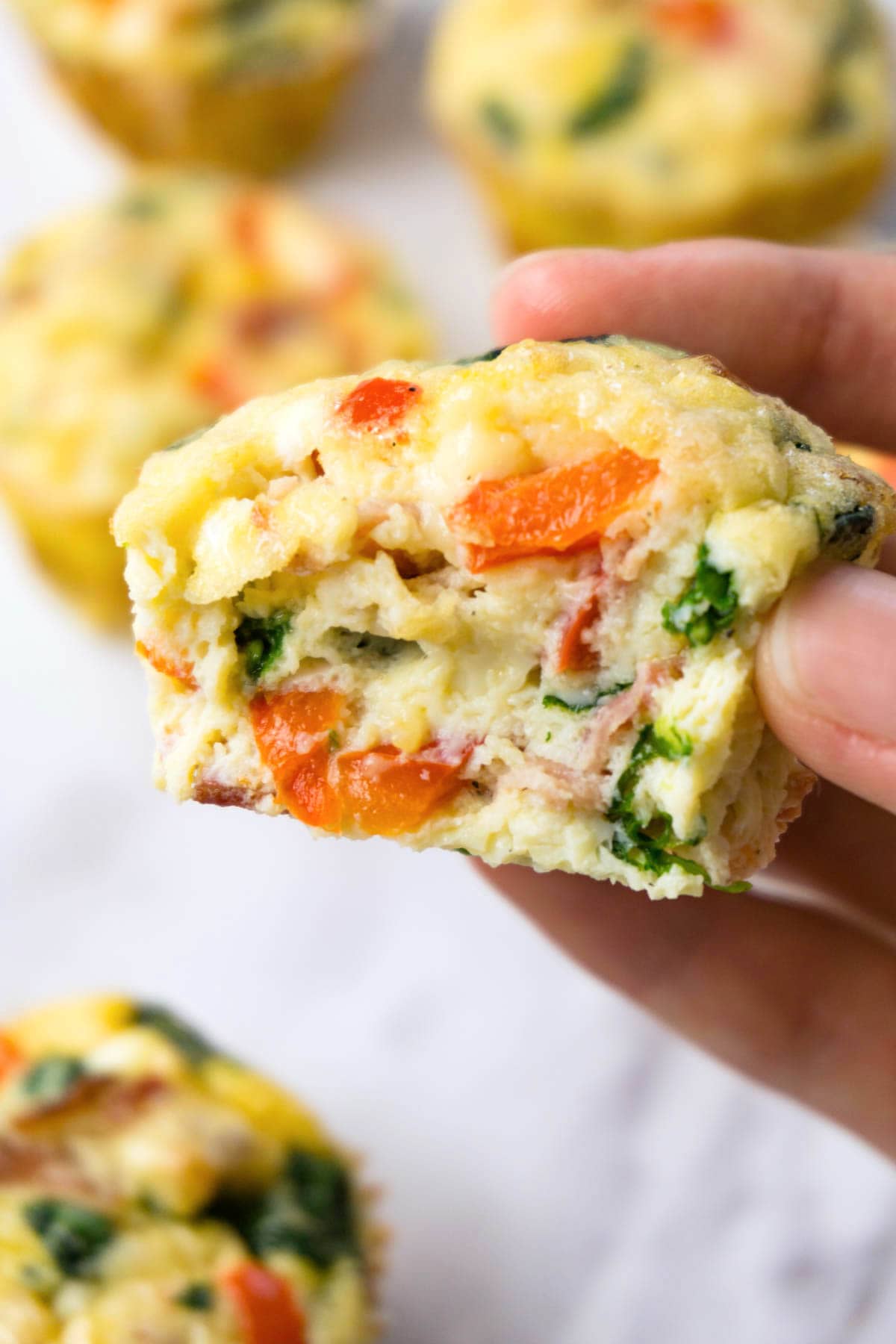 A had holding an egg muffin with spinach, red bell pepper and bacon bits, one bite taken.