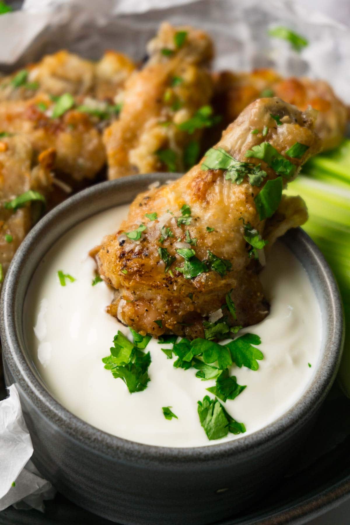 Baked chicken drumette garnished with chopped cilantro in a bowl with creamy dipping sauce, more wings and drumettes on the background.