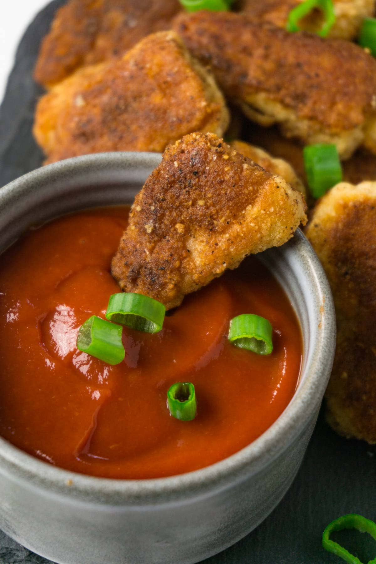 One chicken nugget in a small bowl with ketchup, more nuggets on the background.