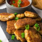 Chicken nuggets on a black board garnished with chipped green onions, a bowl with ketchup on the background.