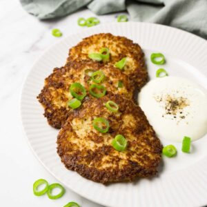 A white round plate with 3 cauliflower hash brown patties served with with chopped green onions and sour cream.