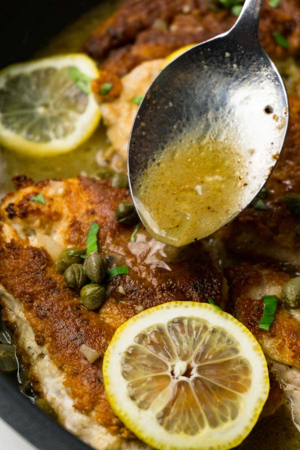 Chicken piccata on a skillet garnished with fresh lemon slices and cappers, a spoon pouring the piccata sauce on the chicken.