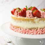 Close up shot of a strawberry cake decorated with pink frosting and fresh strawberries on a white cake stand.