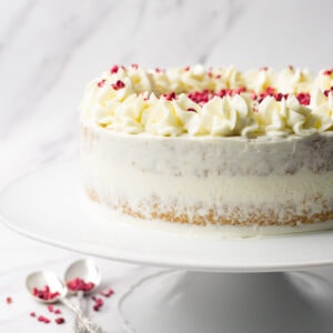 Vanilla caramel cake frosted with cream cheese frosting and decorated with freeze-dried raspberries on a white cake platter.