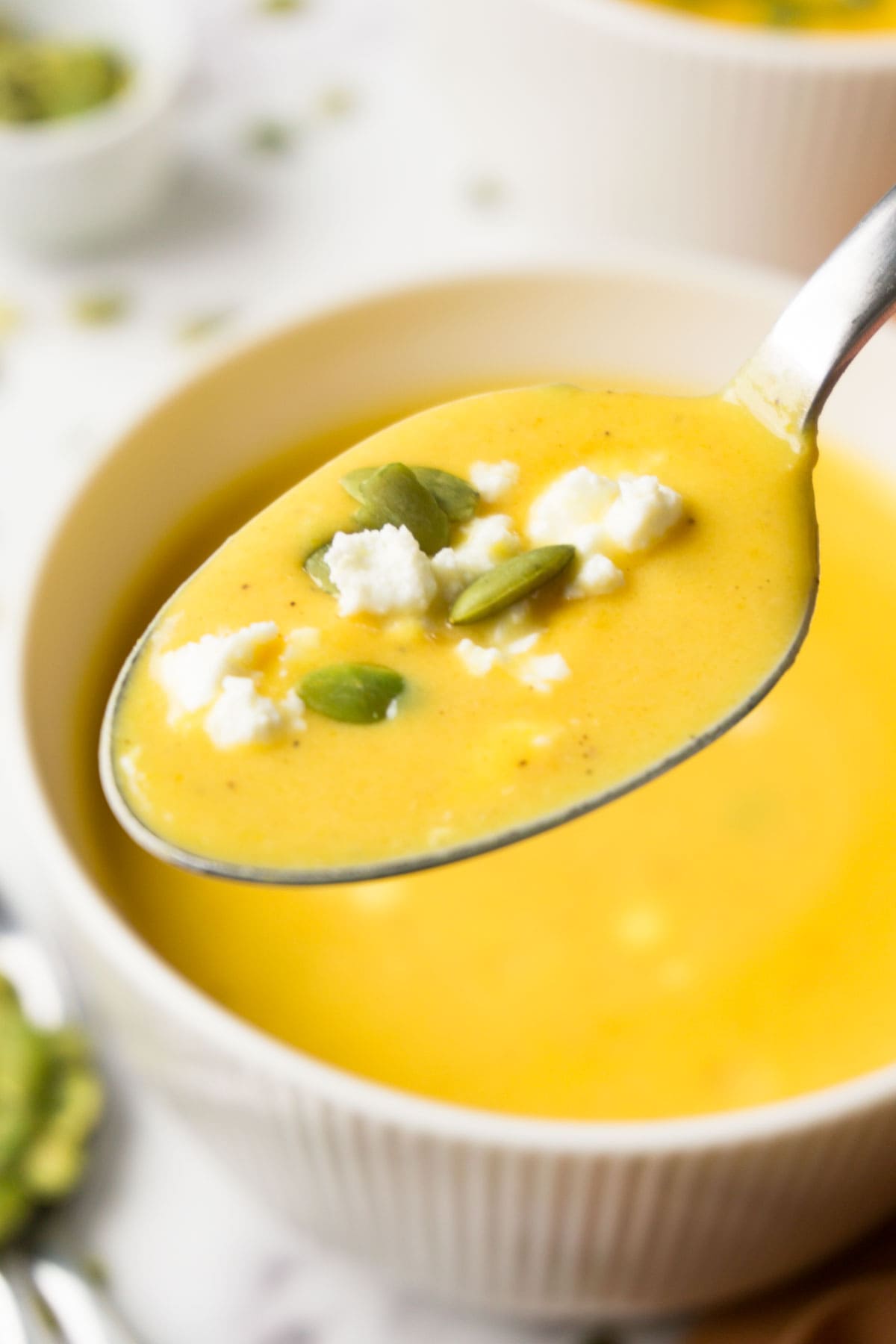 A spoon with butternut squash soup garnished with pepitas and crumbles feta cheese.