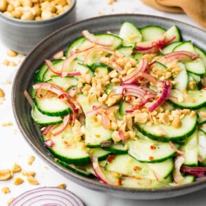 A grey bowl filled with cucumber salad with red onions, red chili pepper flakes and crushed salted peanuts.