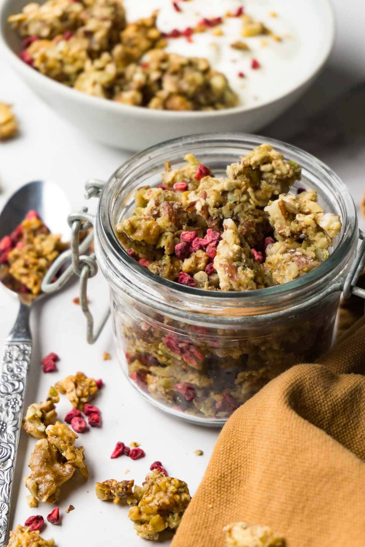 A glass jar full of low carb granola with freeze-dried raspberries, a bowl with yoghurt and granola on the background.