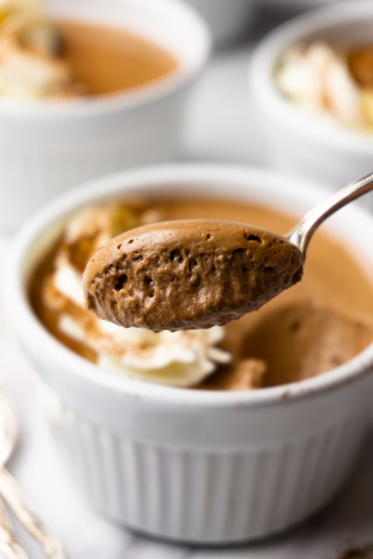 A silver spoon with chocolate mousse, ramekin with the mousse on the background.