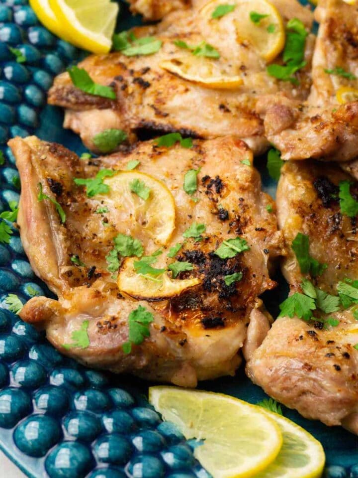 Baked boneless skin-on chicken quarters with thin lemon slices garnished with freshly-chopped cilantro on a turquoise plate.