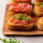 Two pieces of roasted bread with fresh tomato spread. One piece is topped with fresh chives, and another with Jamón Serrano.
