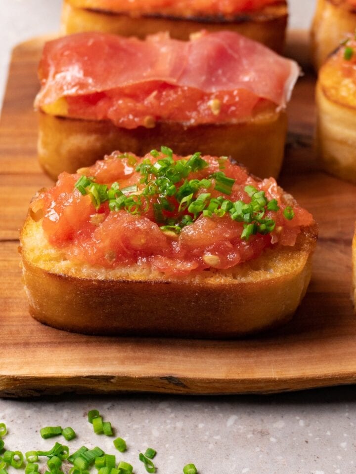 Two pieces of roasted bread with fresh tomato spread. One piece is topped with fresh chives, and another with Jamón Serrano.