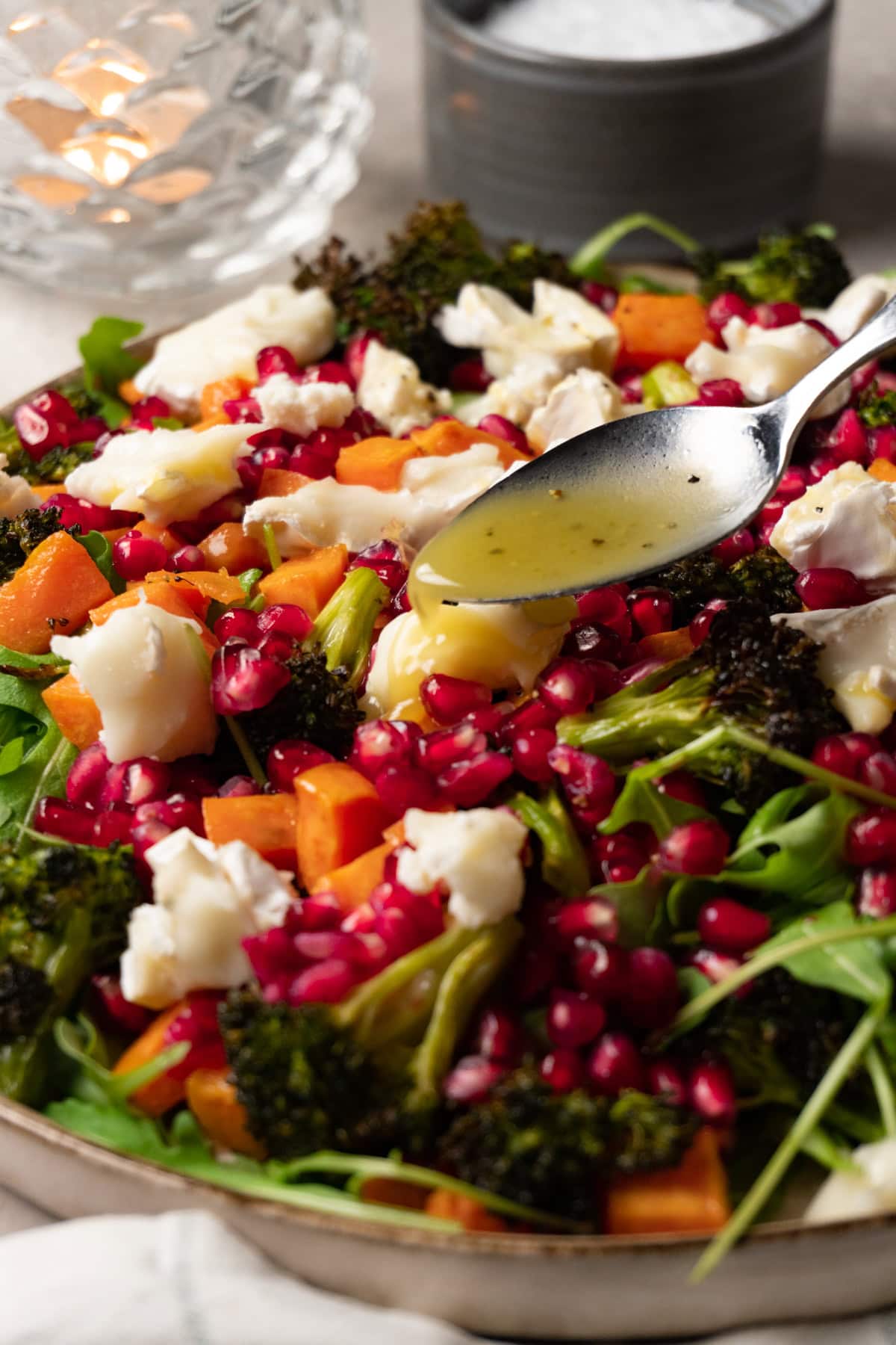 A large plate of salad with roasted sweet potatoes, broccoli, arugula, pomegranate, and goat cheese, with a spoon drizzling dressing on top.